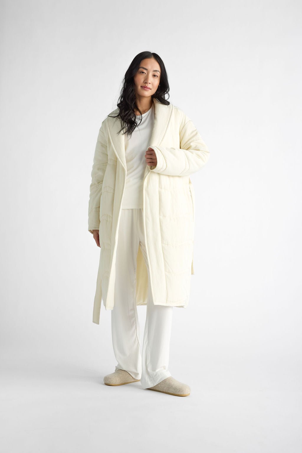 A person is wearing Cozy Earth's Women's Quilted House Coat as part of an all-white set, including pants and slippers, against a plain white backdrop. The individual has long dark hair and looks relaxed. |Color:Vanilla