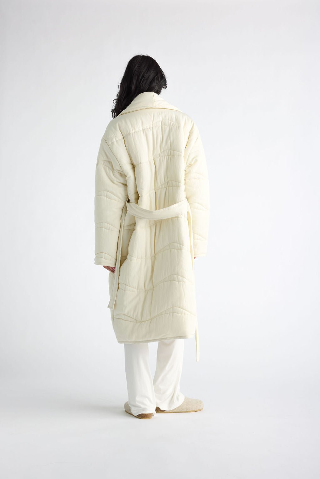 A person with long dark hair, facing away, is wearing Cozy Earth's Women's Quilted House Coat in off-white with a belt and matching wide-legged pants against a plain white studio backdrop.  