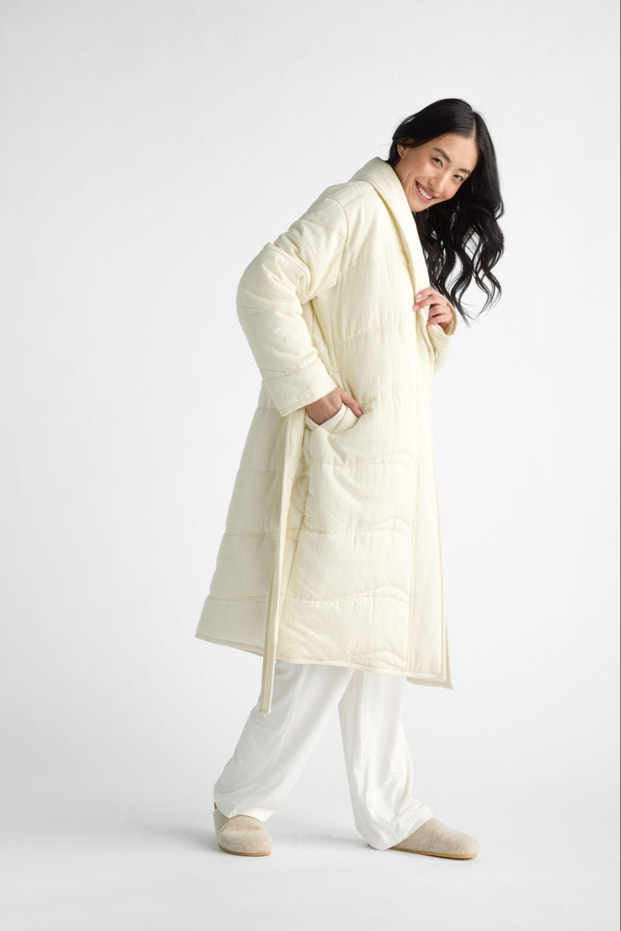 A person models Cozy Earth's Women's Quilted House Coat, a cream-colored quilted robe, on a plain white background. They smile with hands in the coat's pockets, wearing white pants and matching slippers. 