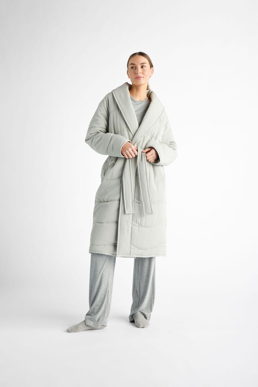 A person against a white background wears a Cozy Earth's Women's Quilted House Coat in light gray over matching loungewear, tied with a belt, and gray socks. |Color:Aloe