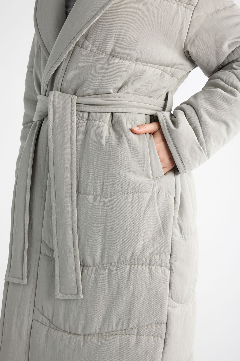 A close-up shows a person in Cozy Earth's Women's Quilted House Coat, light gray and soft-textured, with a relaxed posture. The robe is tied at the waist, one hand in a pocket. 