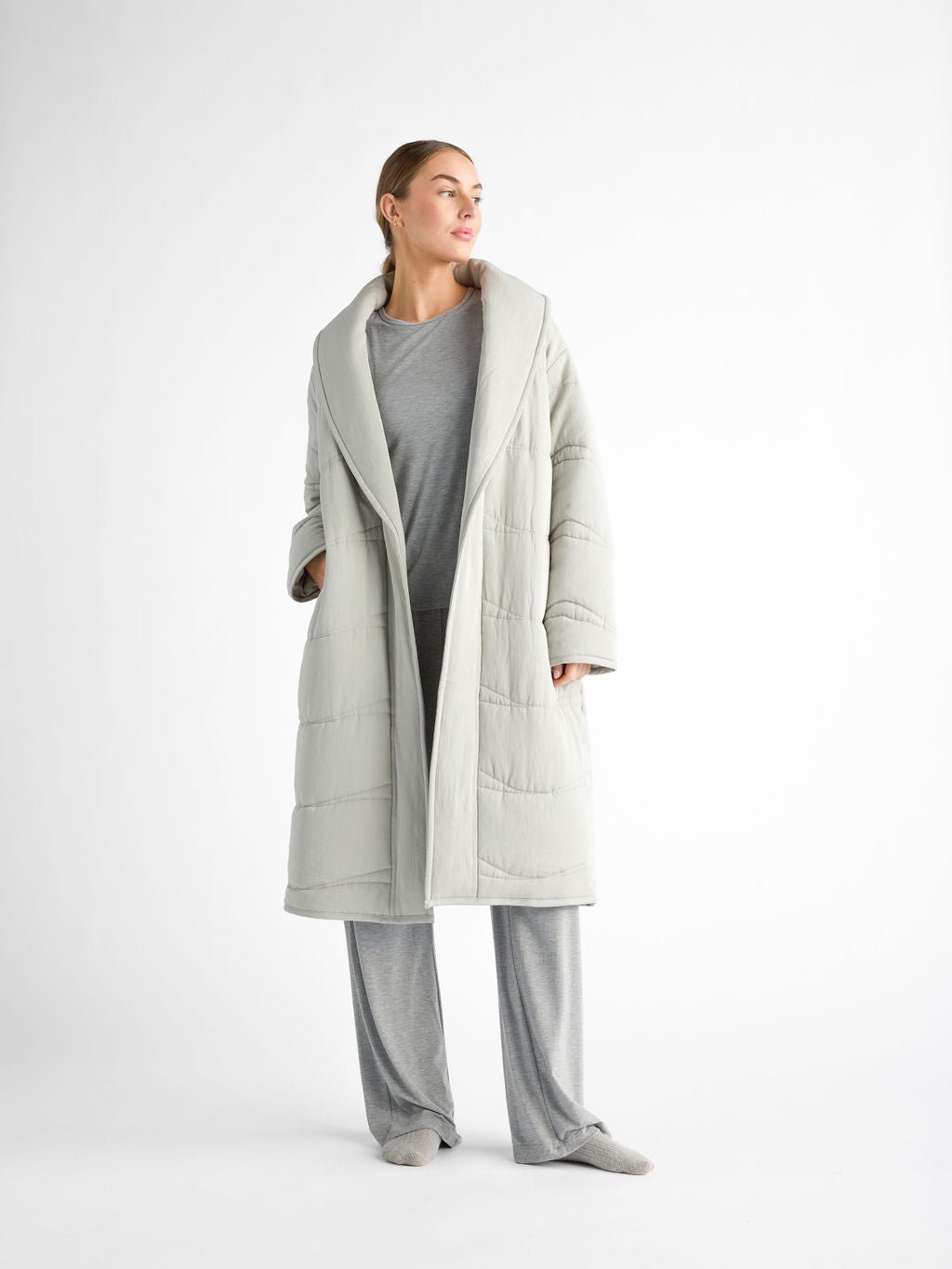 A person in a Cozy Earth's Women's Quilted House Coat over a matching gray loungewear set stands against a plain white background, looking to the side with hands in pockets and wearing socks, creating a relaxed, cozy vibe. 