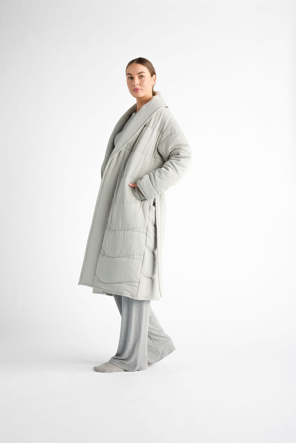 A person is wearing a Cozy Earth Women's Quilted House Coat in light gray, paired with gray pants, standing with hands in pockets against a plain white background. 