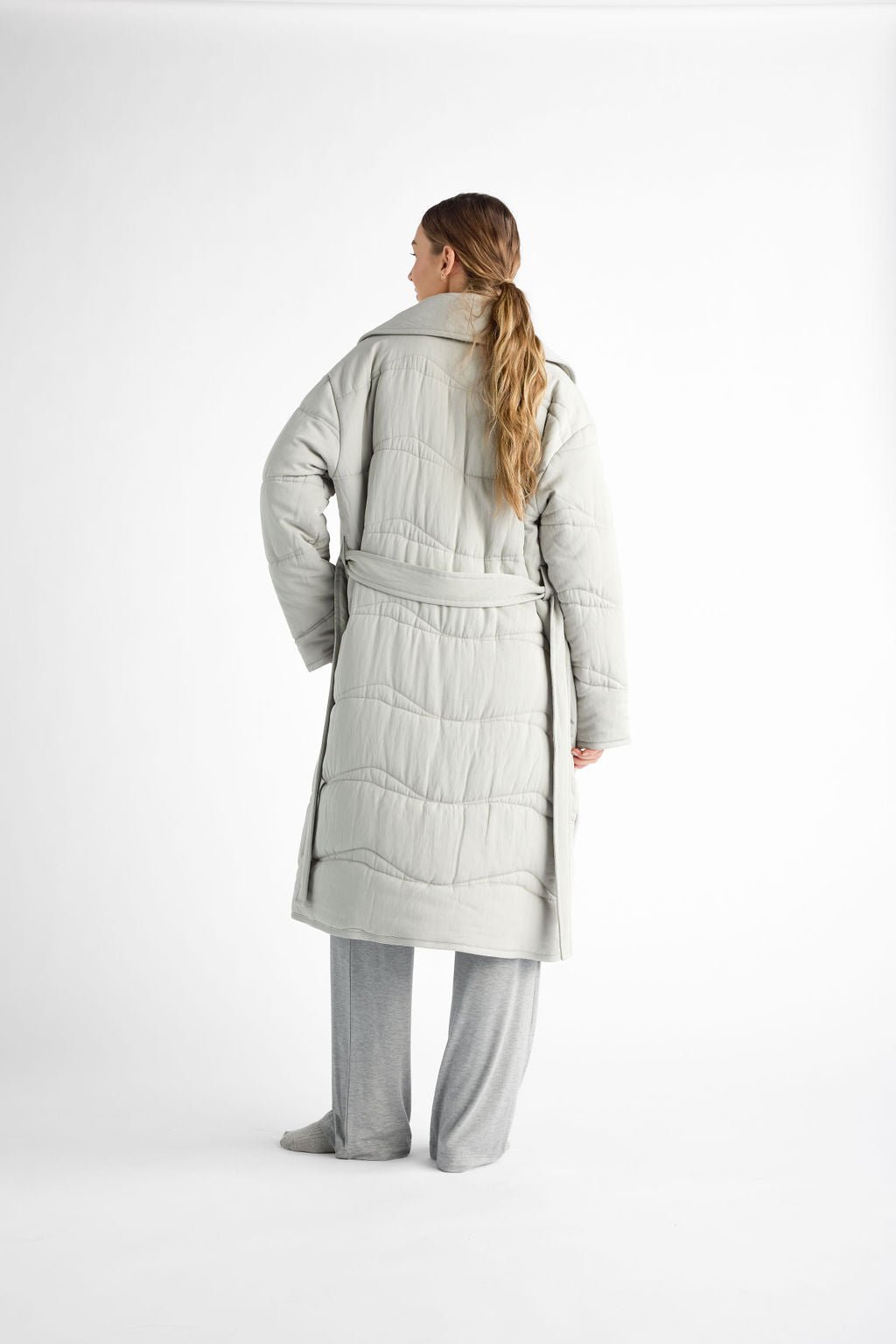 A person with long hair, viewed from the back, wears a light-colored Women's Quilted House Coat by Cozy Earth. They pair it with loose gray pants are barefoot. A plain white background completes the scene. 