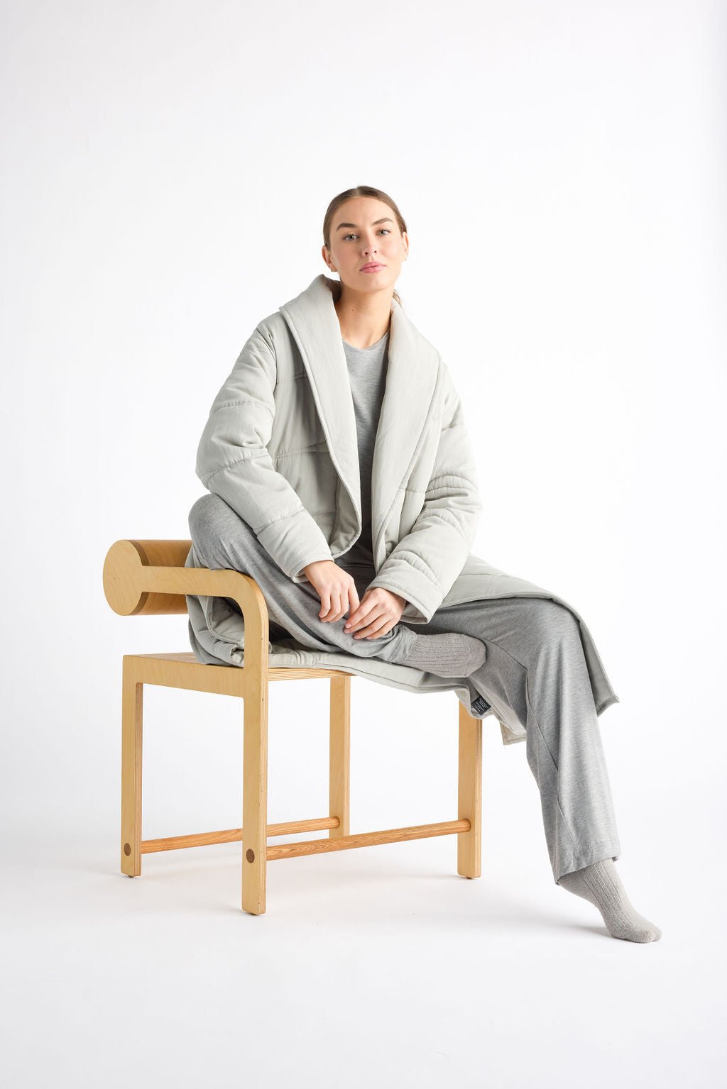 A person wearing a Cozy Earth Women's Quilted House Coat and matching gray pants sits on a wooden chair against a white background. They have short hair and are looking at the camera with a neutral expression. 