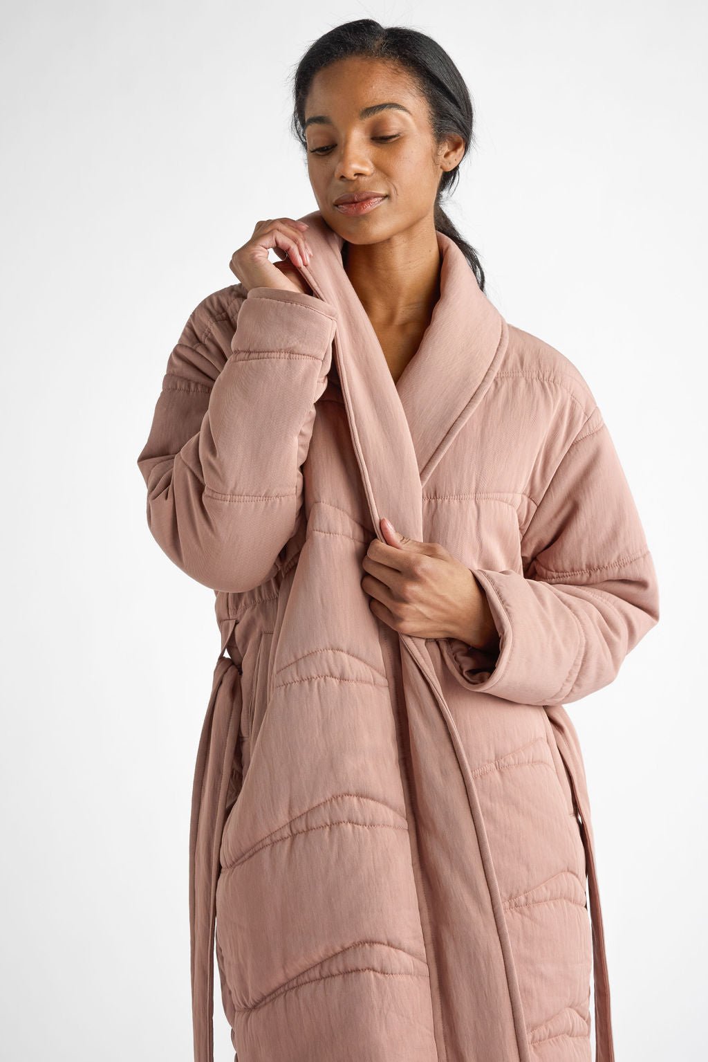 A woman in a Cozy Earth Women's Quilted House Coat, colored pink, stands against a white background. She gently holds the robe's collar with one hand, exuding a soft expression. 