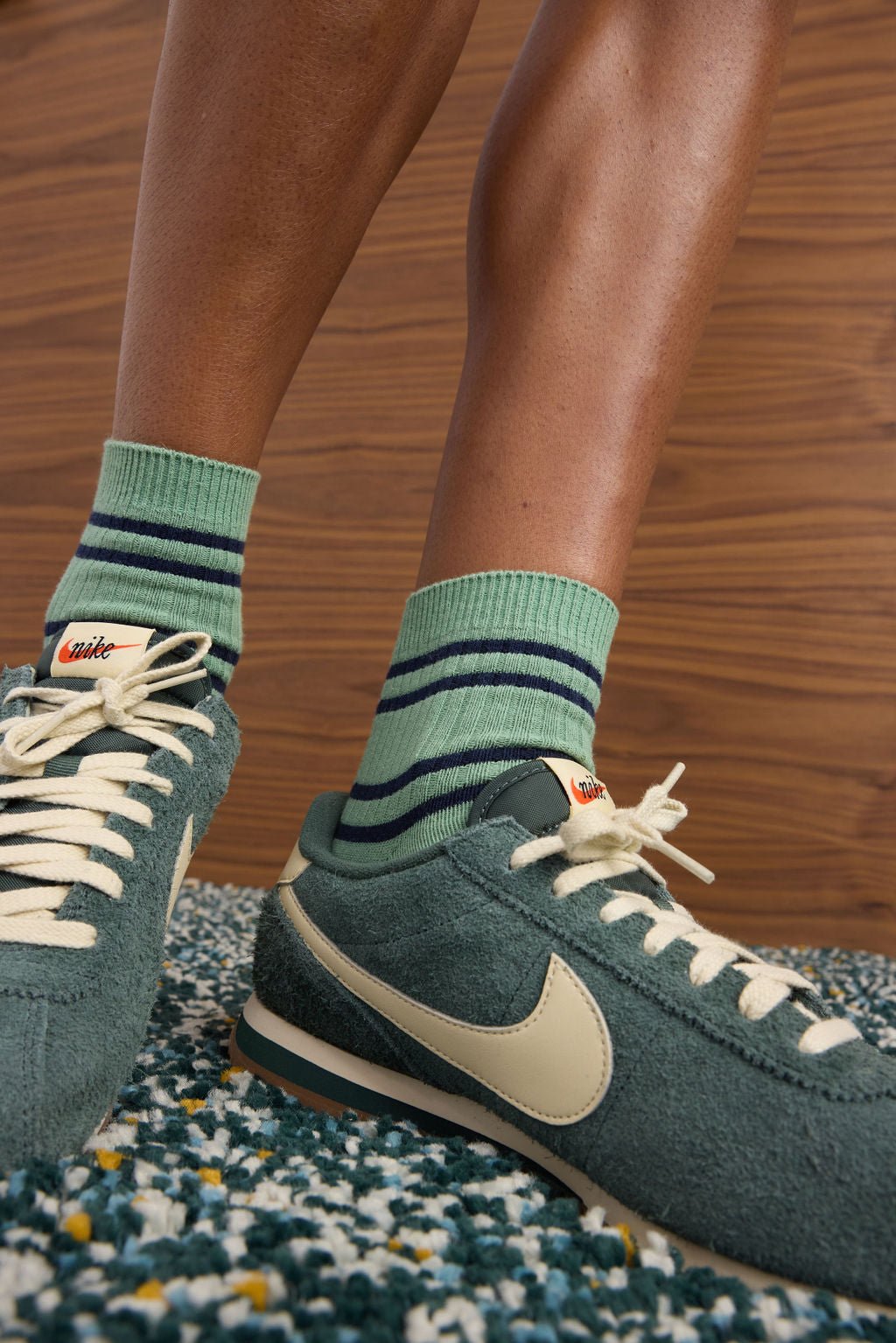 Close-up of a person wearing green Nike sneakers and Cozy Earth's Women's Sadie Sock—green socks with dark blue stripes—while standing on a patterned carpet with a wooden wall in the background. 