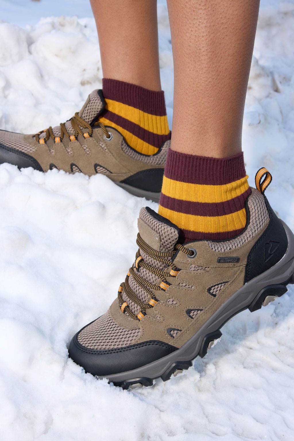 A person stands on snow wearing brown hiking shoes and Cozy Earth Women's Sadie Socks in maroon-and-yellow stripes. 