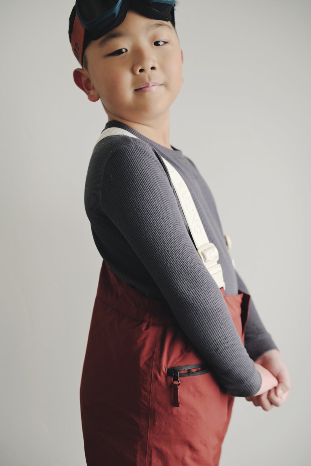 A young child stands confidently against a plain background, wearing the Cozy Earth Children's Bamboo Waffle Knit Base Layer Long Sleeve, red ski pants with suspenders, and ski goggles on their head.