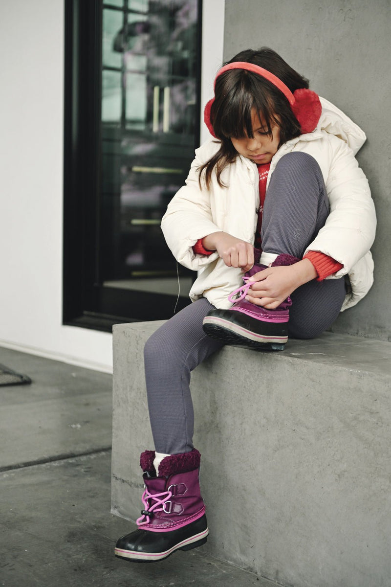 A young girl in Cozy Earth Children's Bamboo Waffle Knit Base Layer Pants sits on a concrete ledge, tying her boot. She also wears a white winter coat, purple boots, gray leggings, and red earmuffs.