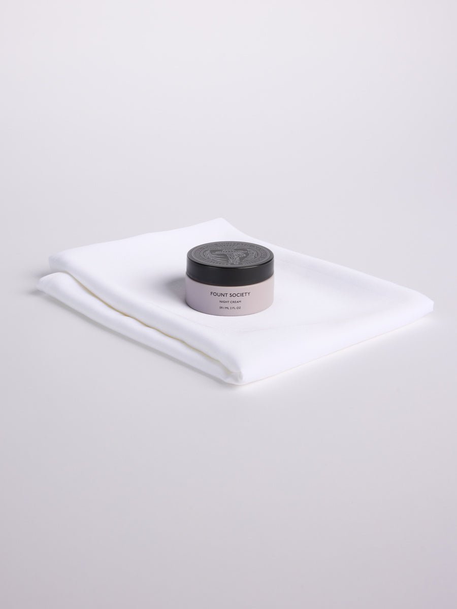 A jar of Fount Society Night Cream sits on a folded set of white Cozy Earth Bamboo Pillowcases.