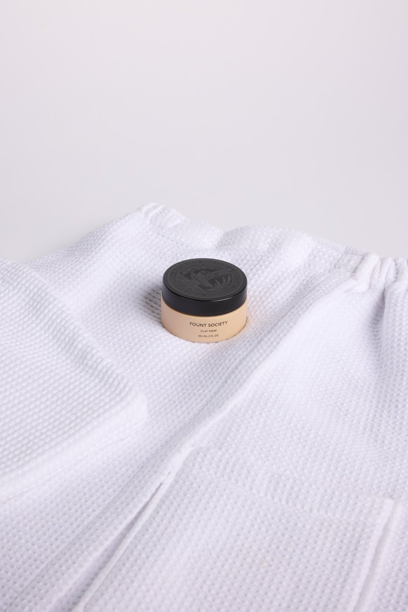 A jar of Fount Society Clay Mask sits on top of a Cozy Earth Waffle Bath Wrap and Waffle Hair Towel on a white background.