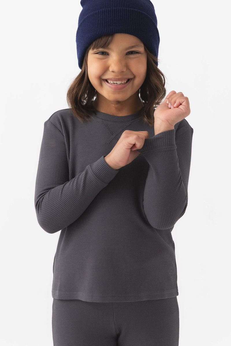 A young child wearing Cozy Earth's Children's Bamboo Waffle Knit Base Layer Long Sleeve in dark gray smiles while holding the sleeve. The beanie is dark blue and the background is plain white.