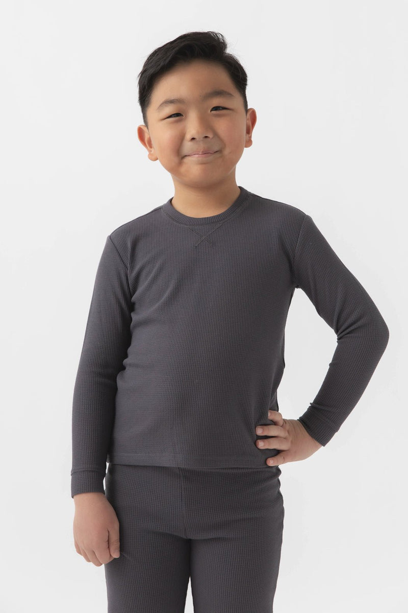 A young boy with short dark hair smiles, standing with one hand on his hip, wearing the Cozy Earth Children's Bamboo Waffle Knit Base Layer Long Sleeve and matching bottoms against a plain white background.