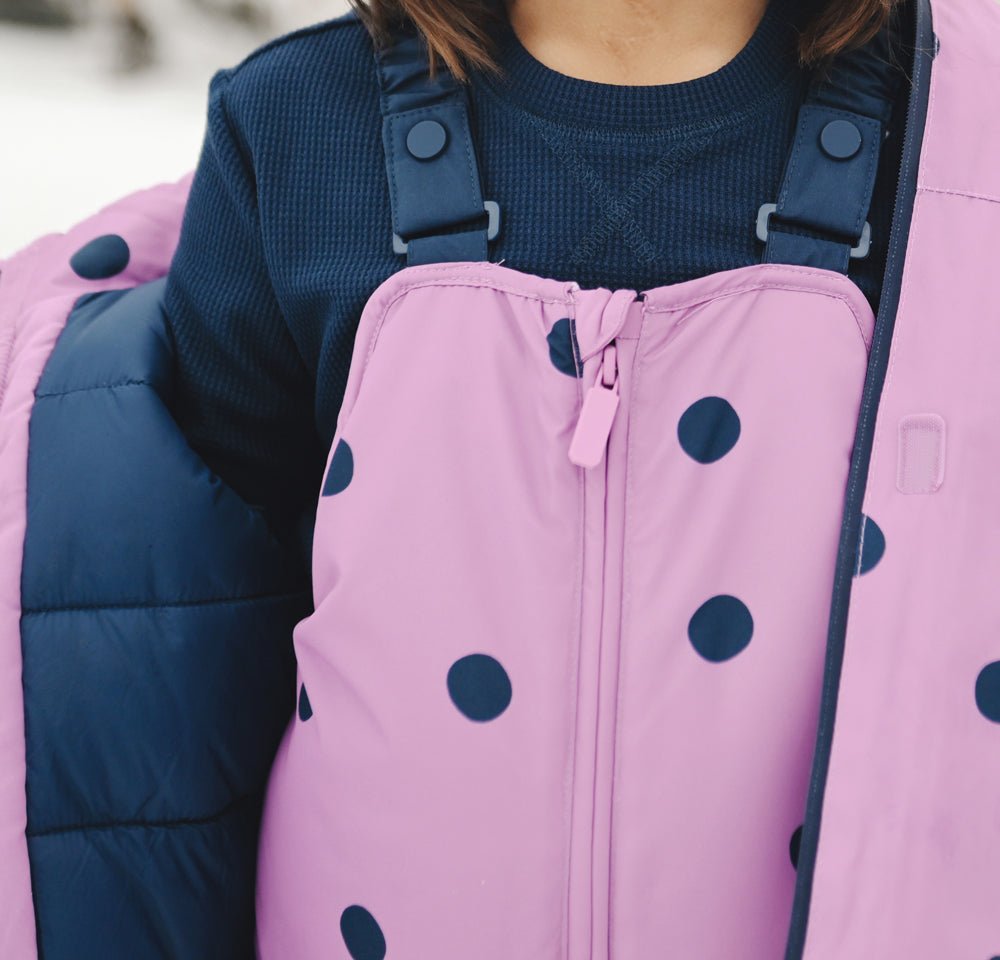 A child wearing the Cozy Earth Children's Bamboo Waffle Knit Base Layer Long Sleeve, paired with a pink overall with large navy blue polka dots and partially covered by a navy puffer jacket. The child’s face is not visible.