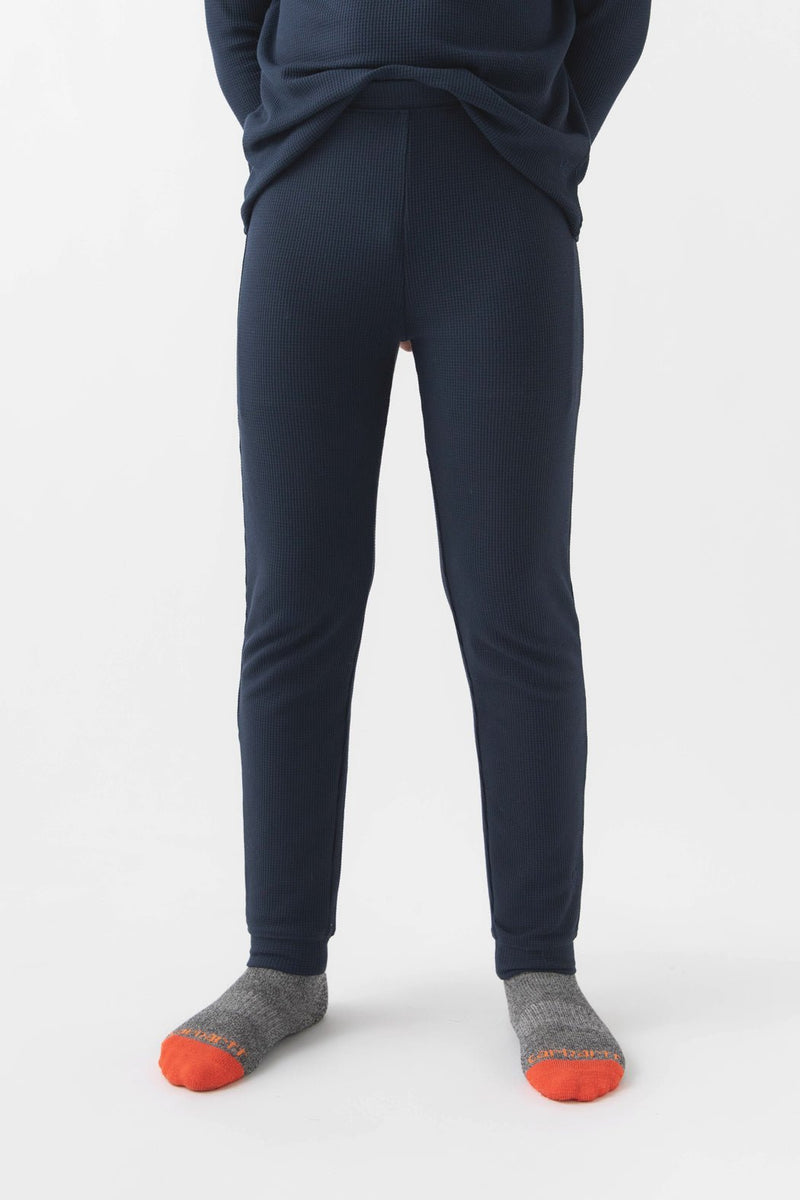 A child wears Cozy Earth Children's Bamboo Waffle Knit Base Layer Pant in navy, paired with gray socks with orange toes, standing against a plain white background. The upper body is mostly out of the frame.
