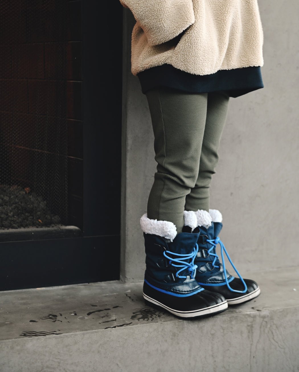 A child in Cozy Earth's Children's Bamboo Waffle Knit Base Layer Pant stands indoors near a fireplace, paired with a beige fleece jacket and blue winter boots with white fur trim on a concrete floor.|Color:Olive