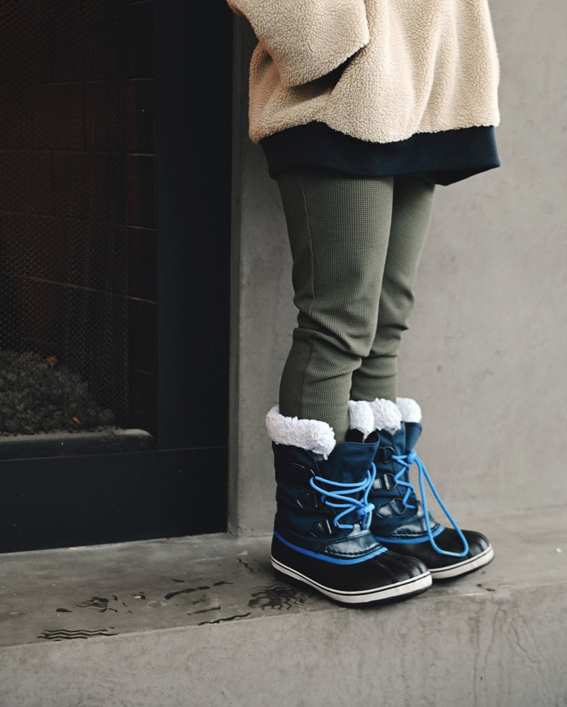 A child in Cozy Earth's Children's Bamboo Waffle Knit Base Layer Pant stands indoors near a fireplace, paired with a beige fleece jacket and blue winter boots with white fur trim on a concrete floor.