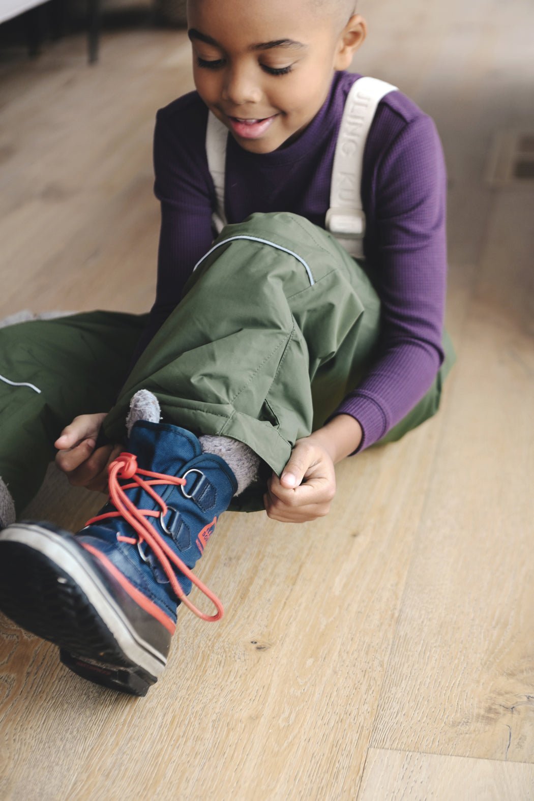 A child wearing green pants and a Cozy Earth Children's Bamboo Waffle Knit Base Layer Long Sleeve smiles while sitting on a wooden floor and putting on blue hiking boots with red laces.