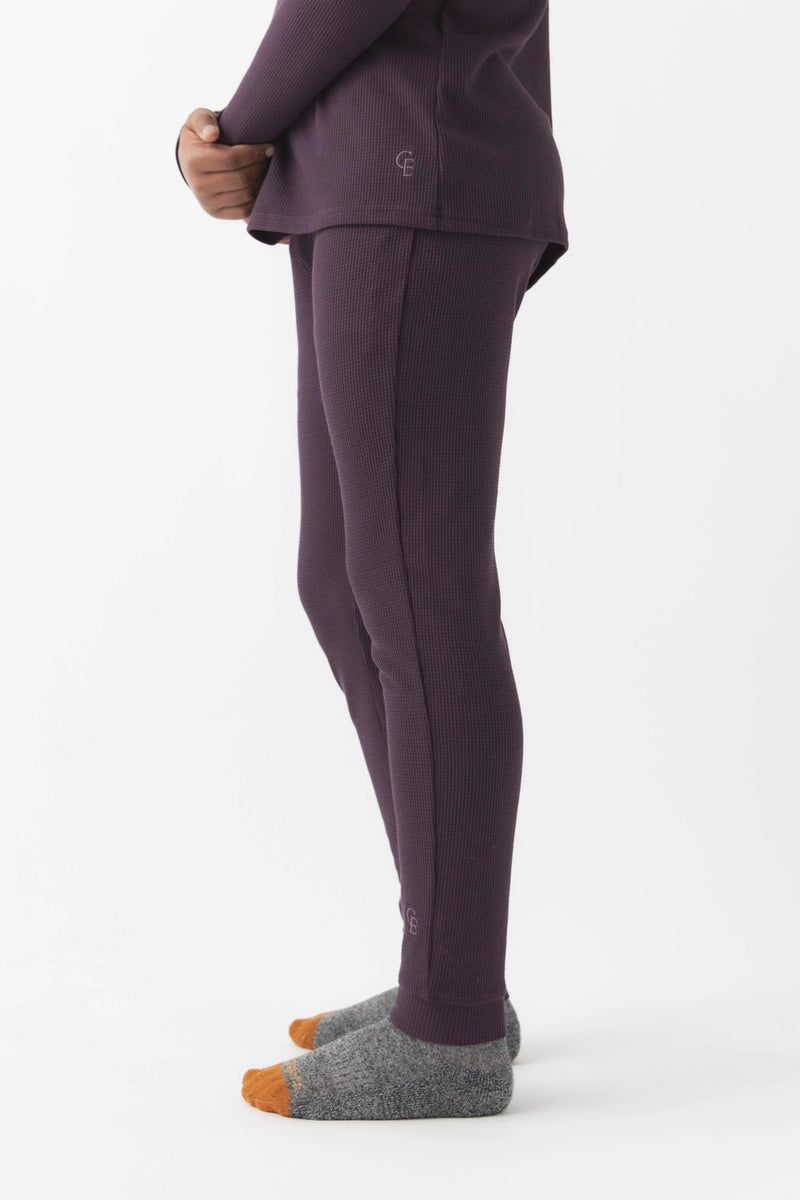 A child stands sideways in Cozy Earth Children's Bamboo Waffle Knit Base Layer Pant, paired with a matching purple thermal top and grey-orange socks, against a plain white background.