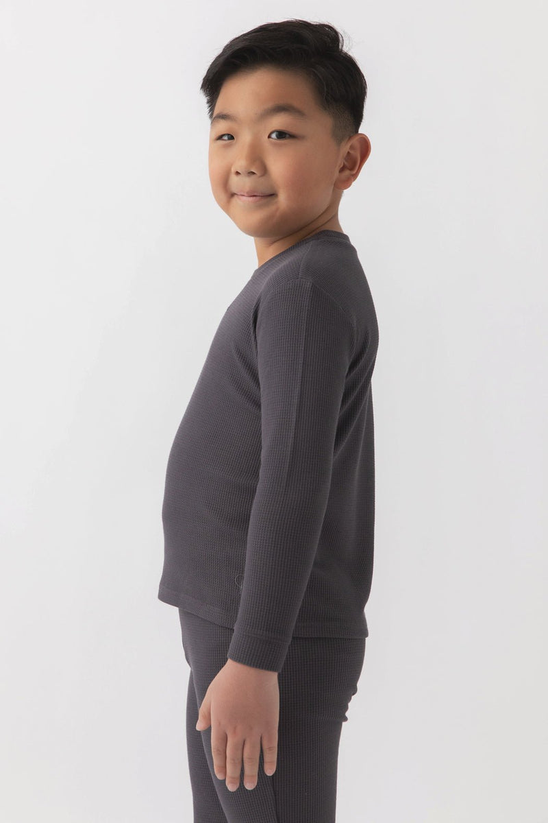 A young boy with short black hair stands sideways, smiling, and wearing the Cozy Earth Children's Bamboo Waffle Knit Base Layer Long Sleeve in dark gray. The background is plain white.