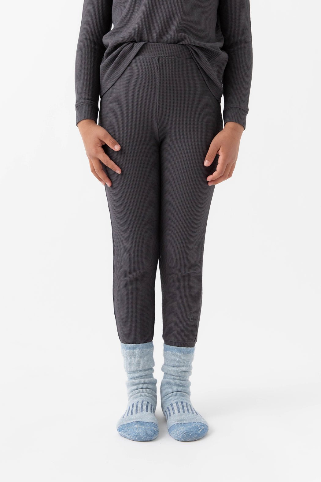 A child stands on a white background wearing Cozy Earth's Children's Bamboo Waffle Knit Base Layer Pant in Iron, with matching long-sleeve top and light blue patterned socks, hands by their sides.|Color:Iron