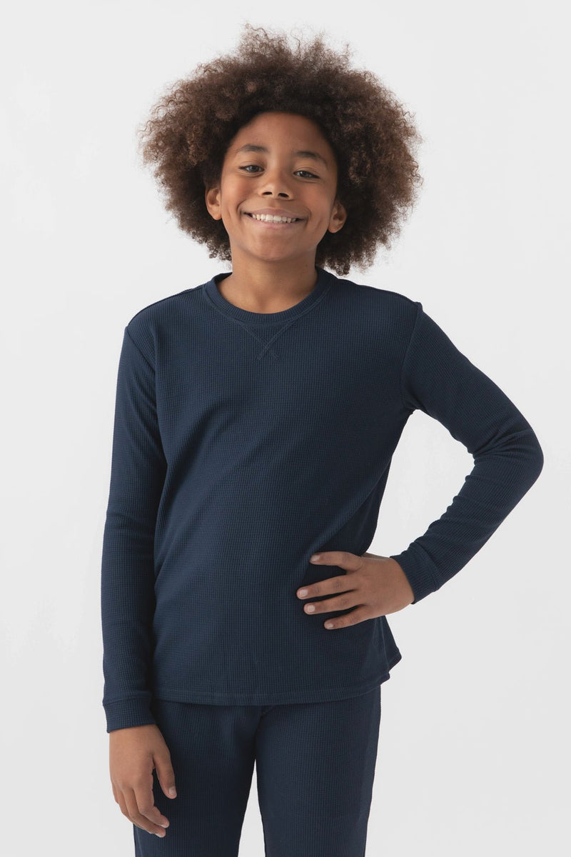 Smiling child with curly hair wears Cozy Earth's Children’s Bamboo Waffle Knit Base Layer Long Sleeve in navy blue, standing against a plain white background with one hand on their hip.