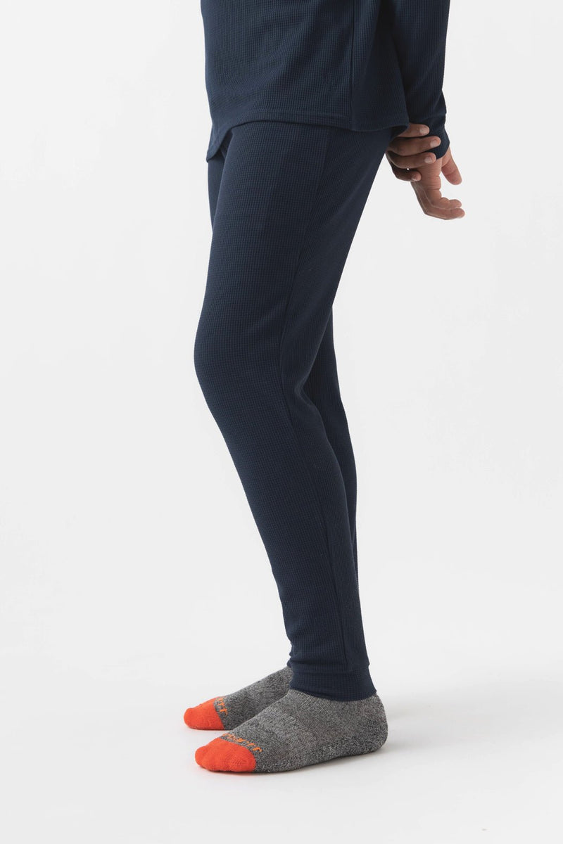 A child’s lower body is shown on a white background, wearing Cozy Earth Children’s Bamboo Waffle Knit Base Layer Pants in navy, a matching long-sleeve top, and gray socks with bright orange toes and heels.