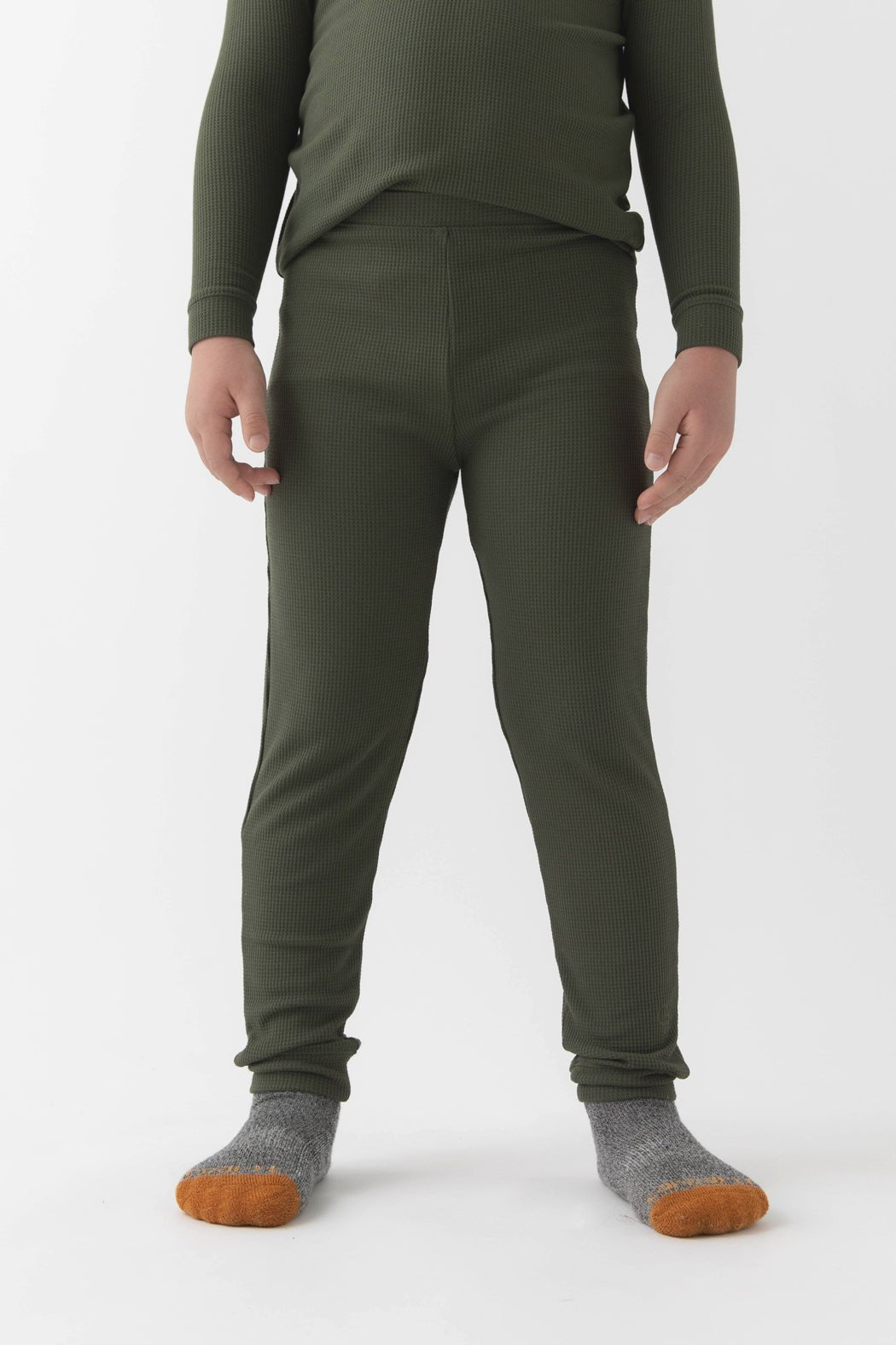 A child wears Cozy Earth’s Children’s Bamboo Waffle Knit Base Layer Pant in Olive, paired with a matching long-sleeve top and gray socks with orange toes, standing against a plain white background. The child’s face is not shown.