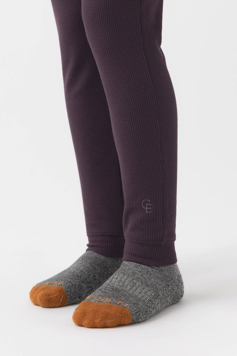 A child in Cozy Earth Children's Bamboo Waffle Knit Base Layer Pants, featuring a textured dark purple look, pairs them with gray socks with orange toes, standing on a white background.
