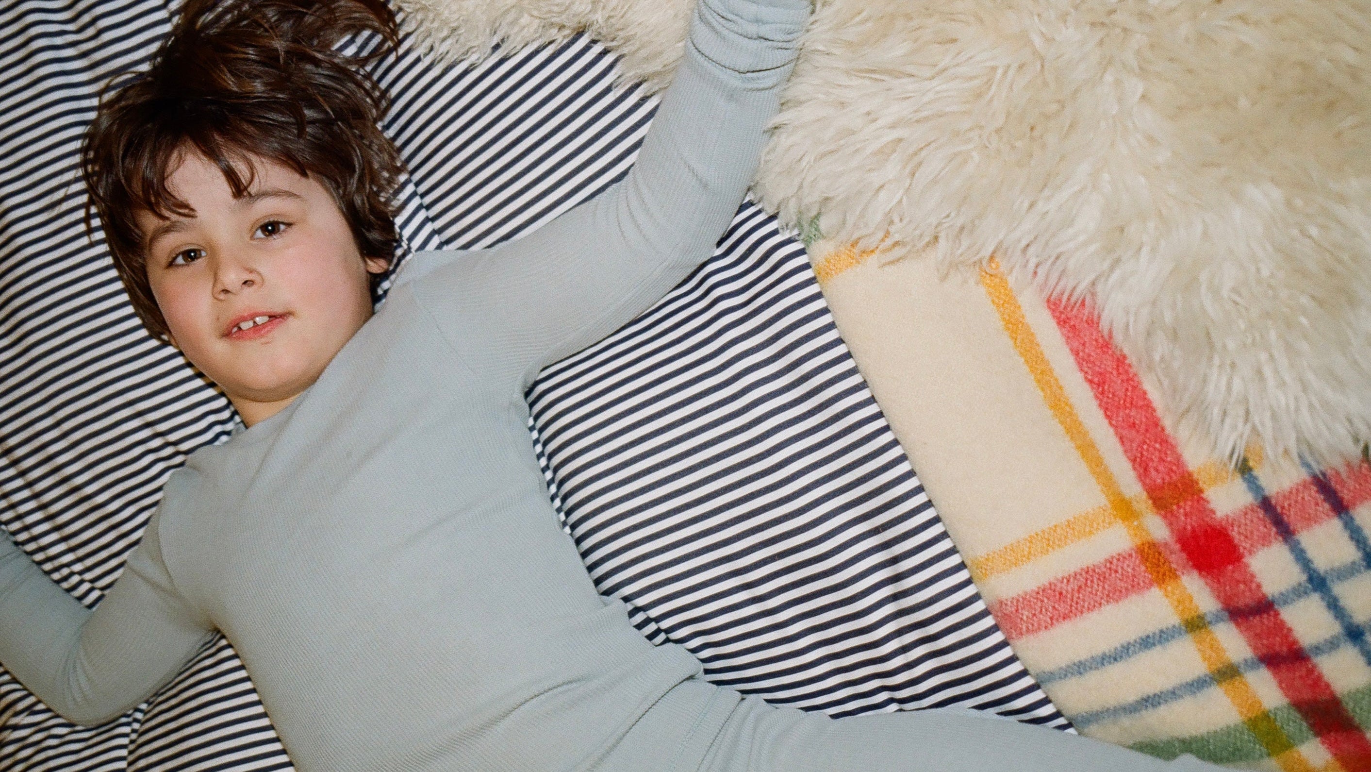 Child in light gray pajamas lying on a striped pillow with a plaid blanket