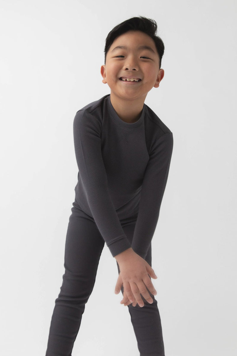 A young boy with short black hair smiles while leaning forward, wearing the Cozy Earth Children's Bamboo Waffle Knit Base Layer Long Sleeve and matching pants against a plain white background.