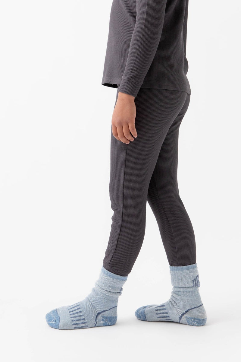 A child wears Cozy Earth's Children's Bamboo Waffle Knit Base Layer Pant in dark gray with a matching ribbed top and light blue socks, standing sideways against a plain white background.