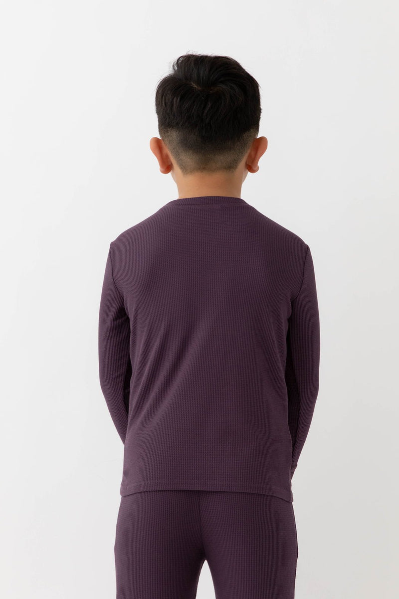 A child with short dark hair stands with their back to the camera, wearing Cozy Earth’s Children’s Bamboo Waffle Knit Base Layer Long Sleeve in purple, paired with matching pants, against a plain white background.