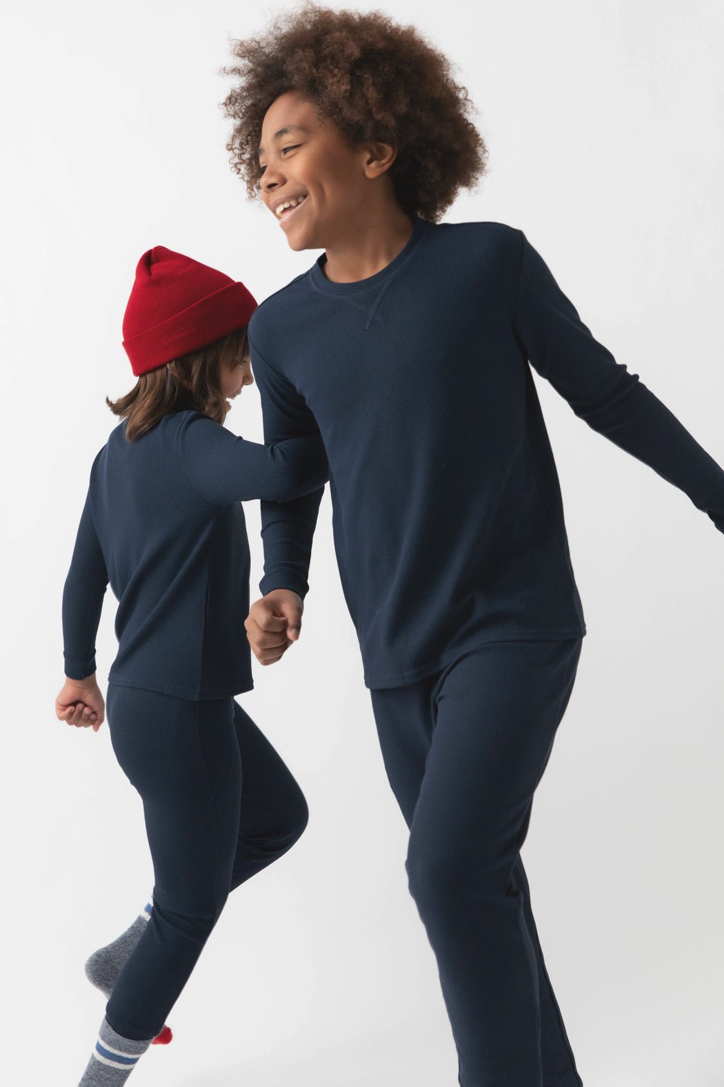 Two children wearing matching Cozy Earth Children's Bamboo Waffle Knit Base Layer Long Sleeves, one with a red beanie, happily holding hands and walking together against a plain white background.
