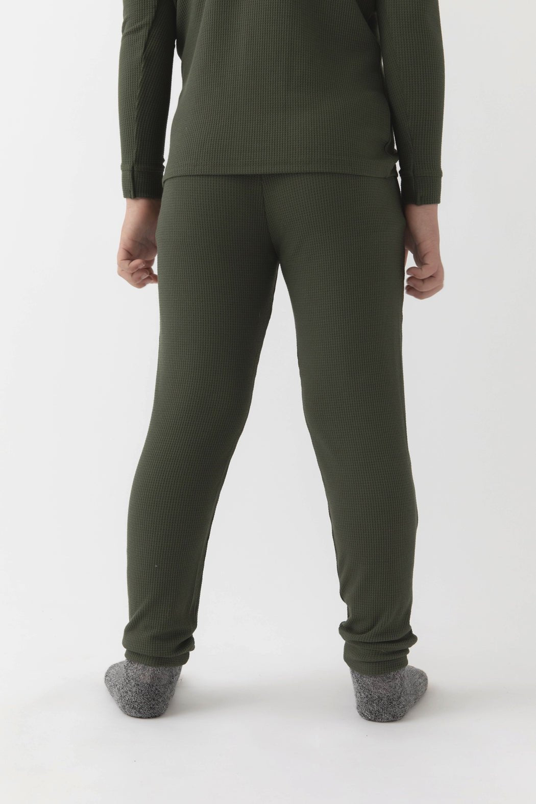 A child faces away from the camera wearing Cozy Earth's Children's Bamboo Waffle Knit Base Layer Pant in olive green, paired with a matching ribbed long-sleeve top and gray socks, set against a plain white background.|Color:Olive