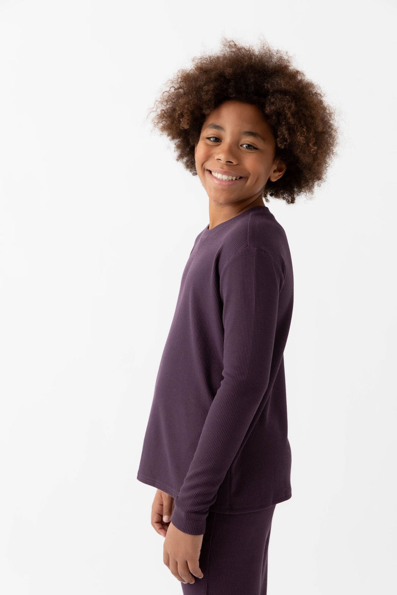 A smiling child with curly hair wears the Cozy Earth Children's Bamboo Waffle Knit Base Layer Long Sleeve in dark purple, paired with matching pants, standing against a plain white background and facing slightly to the side.