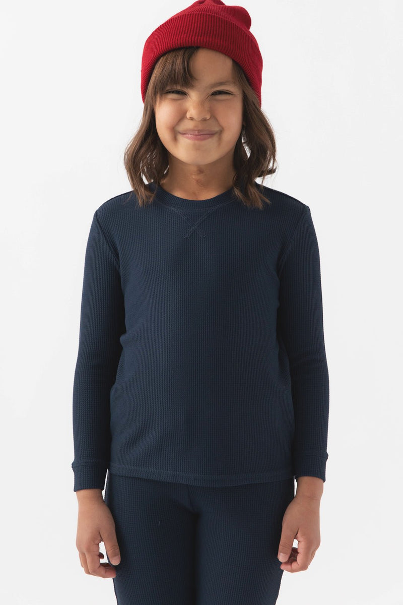 A young child smiles in a red beanie and Cozy Earth's Children's Bamboo Waffle Knit Base Layer Long Sleeve, paired with matching navy blue pants, standing against a plain white background.