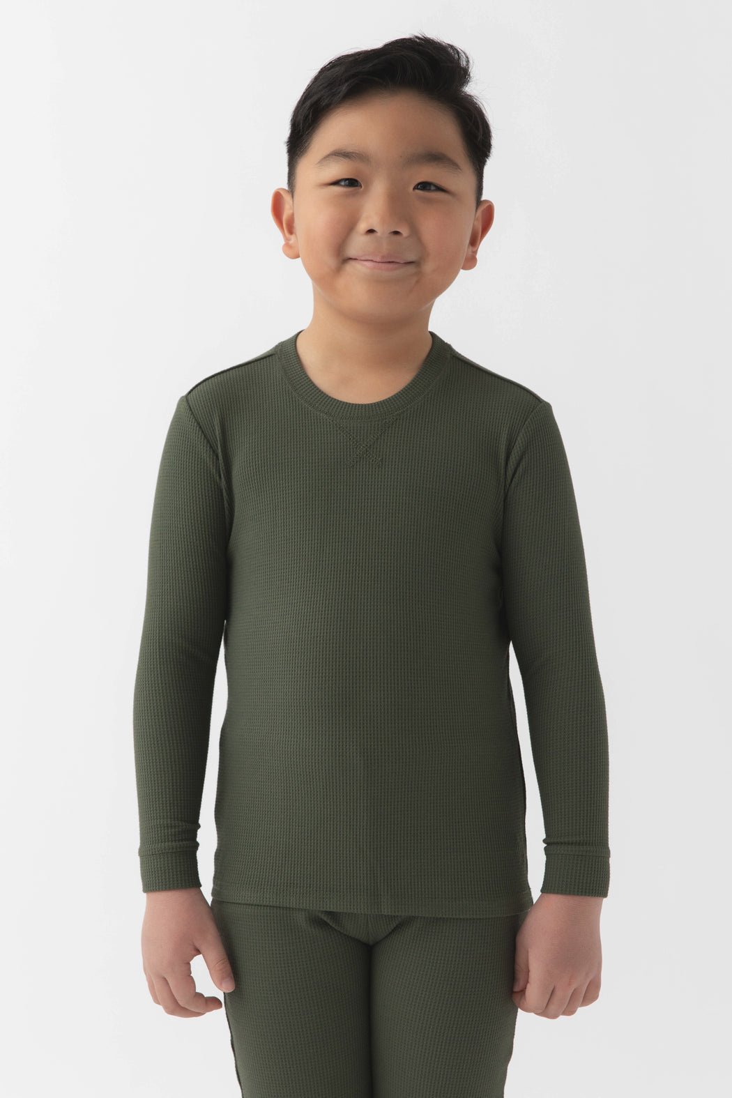 A young boy with short black hair smiles at the camera, wearing the Cozy Earth Children's Bamboo Waffle Knit Base Layer Long Sleeve in olive green. He stands against a plain white background.|Color:Olive