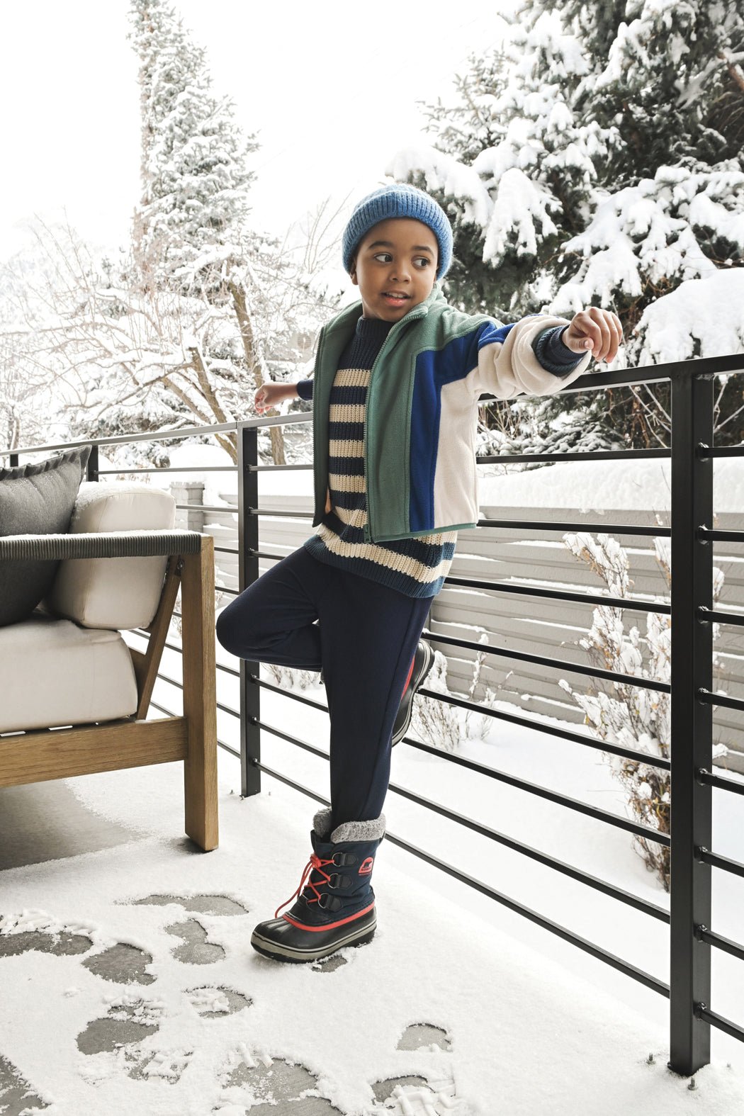 A child smiles and looks to the side on a snowy patio, dressed in Cozy Earth's Children's Bamboo Waffle Knit Base Layer Pant, winter layers, and a blue beanie. Snow-covered trees and furniture appear in the background.
