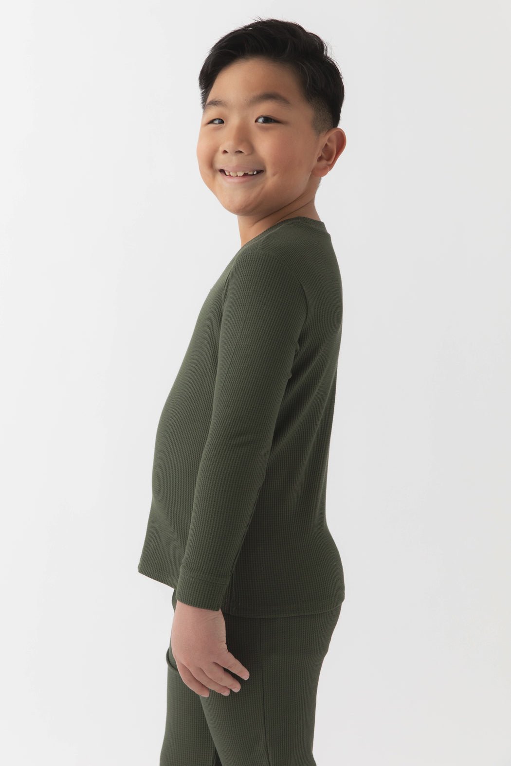 A young boy with short black hair smiles sideways, wearing Cozy Earth's Children's Bamboo Waffle Knit Base Layer Long Sleeve in dark green, paired with matching pants against a plain white background.