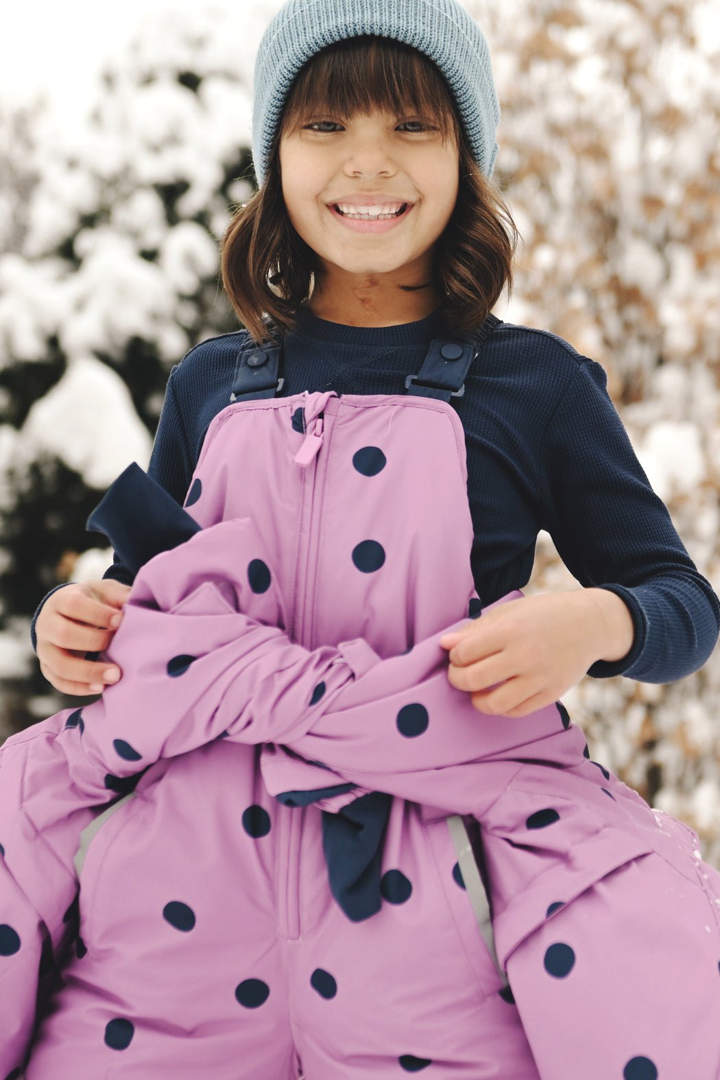 A smiling child wearing the Cozy Earth Children's Bamboo Waffle Knit Base Layer Long Sleeve stands outdoors in snowy surroundings, paired with a blue knit hat and a pink polka-dotted snowsuit.