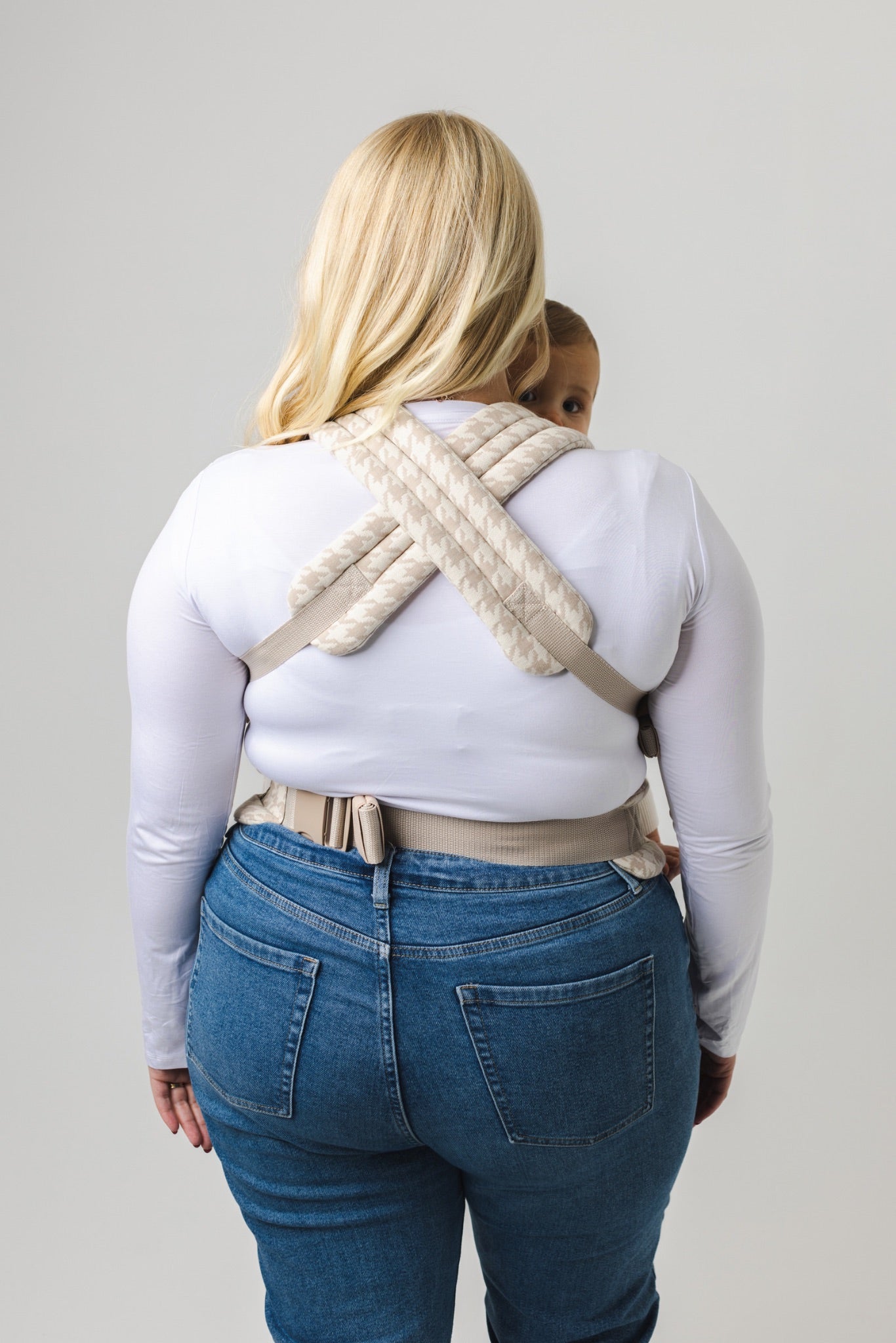 A woman with blonde hair, in a white long-sleeve shirt and blue jeans, carries a baby in the Mabē Monarch Carrier - Houndstooth. The rear-view photo shows the baby peeking over her shoulder.