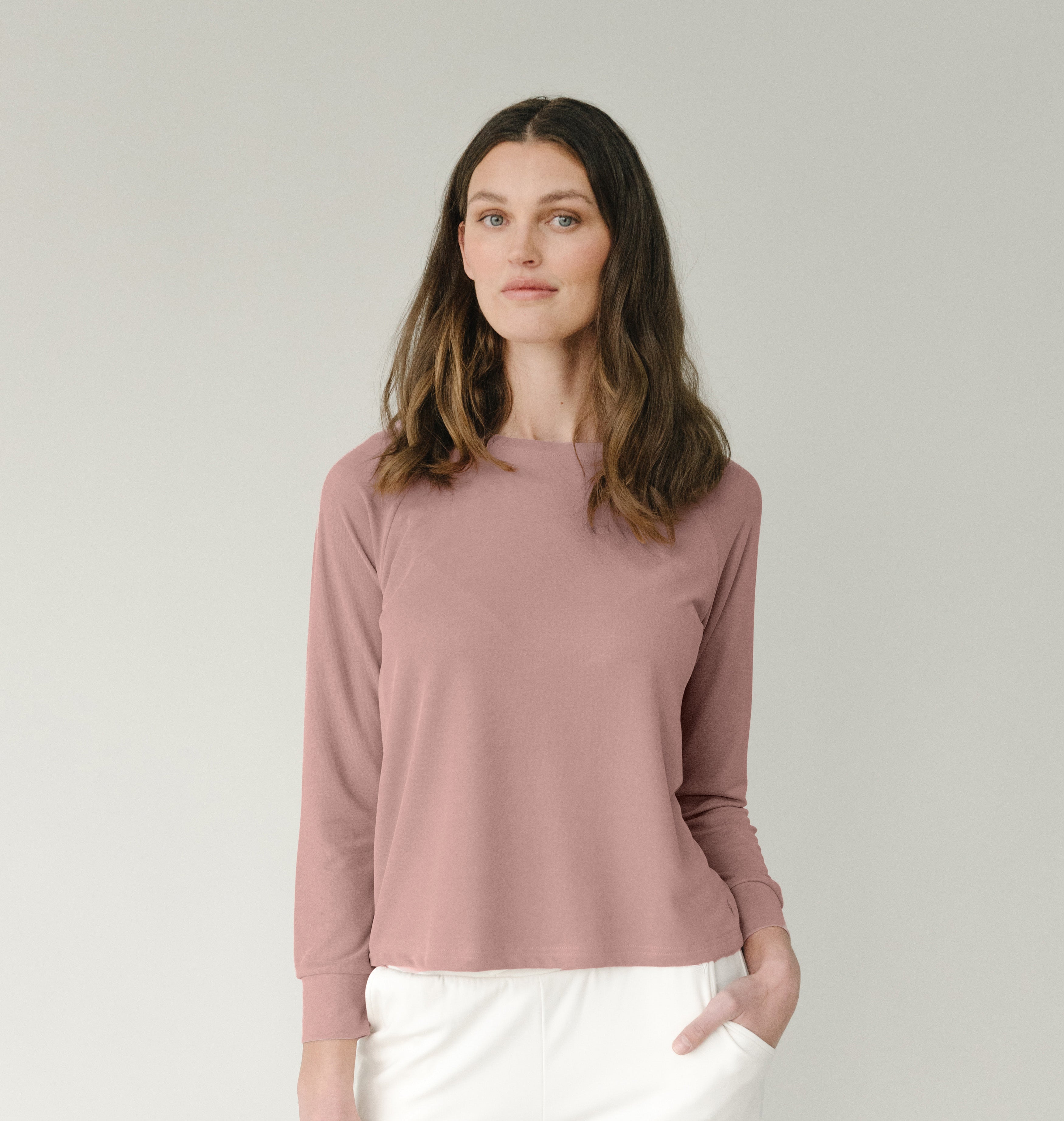 A woman with long brown hair, wearing the Cozy Earth Women's Modern Modal Pullover Crew in pink and white pants, stands against a plain beige background. She has a calm expression and one hand in her pocket.