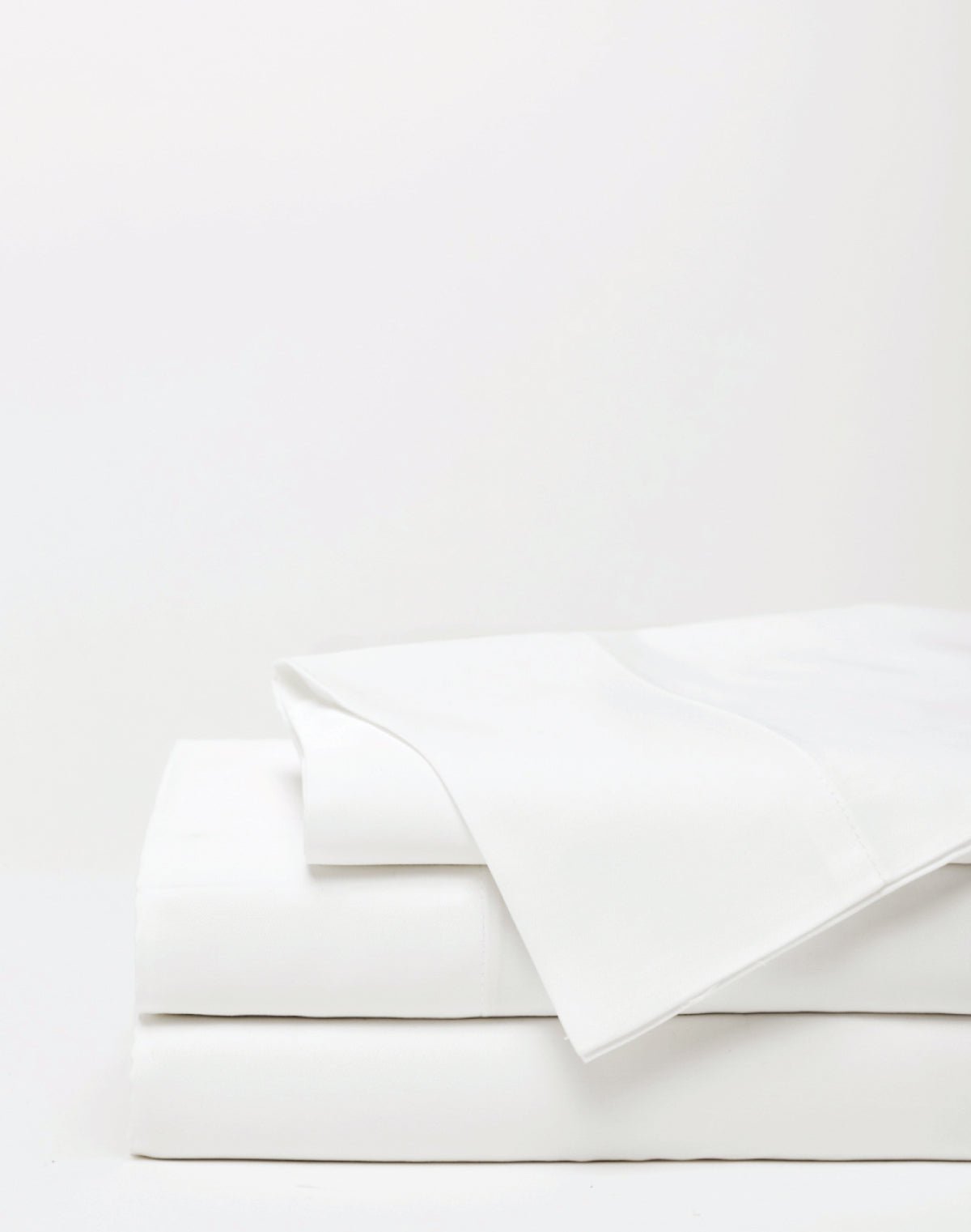 A neatly folded stack of Cozy Earth Bamboo Sheet Set x Loftie Clock bed sheets is displayed on a plain white surface, with one sheet partially draped over the top and a minimal background.