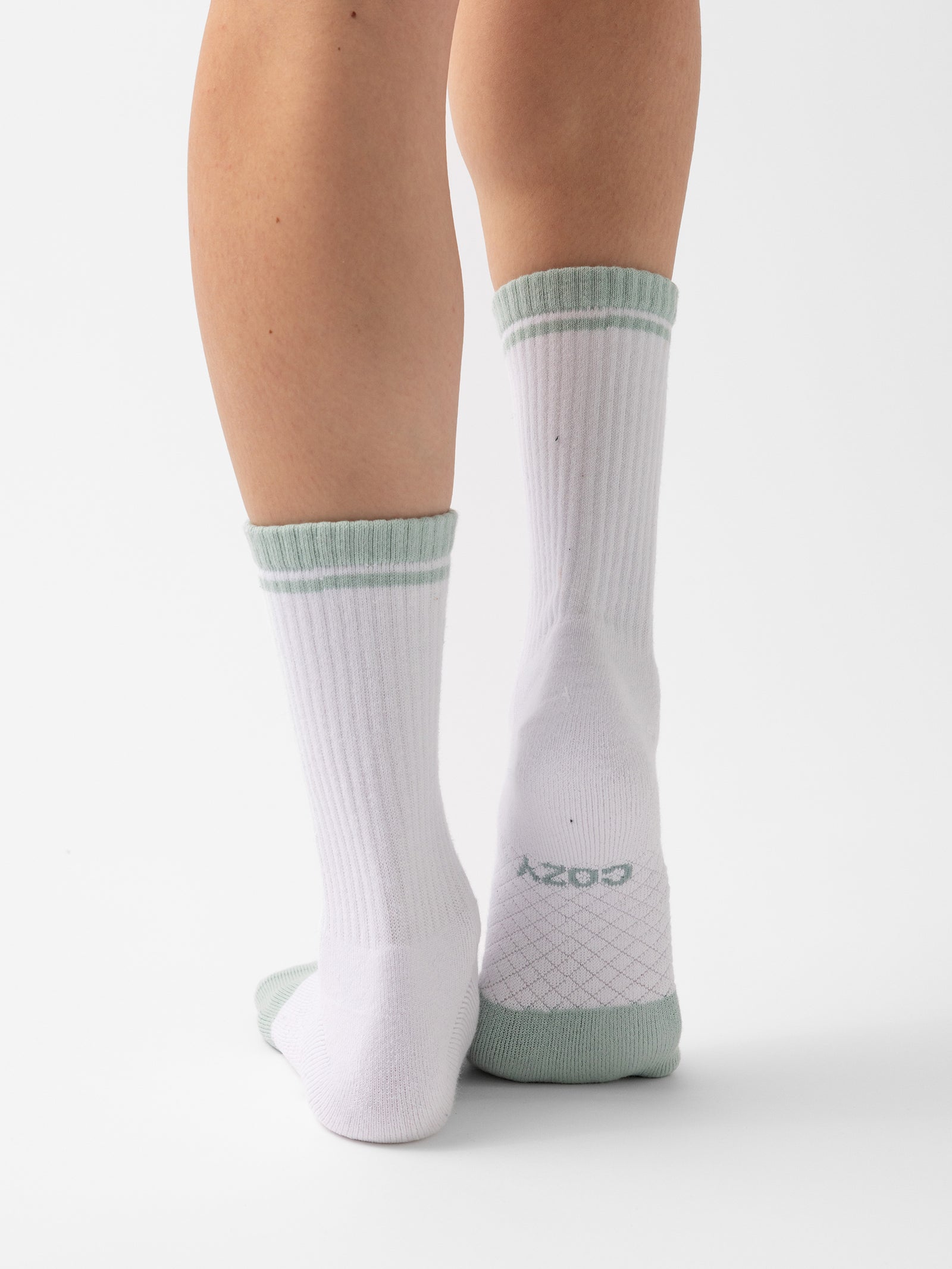 Close-up of a person's lower legs wearing white Essential Calf Socks from Cozy Earth, featuring light green accents at the cuffs, heels, and toes. The socks have vertical ribbing along the legs and a textured sole with the word "cozy" printed on one of them. The background is plain white. 