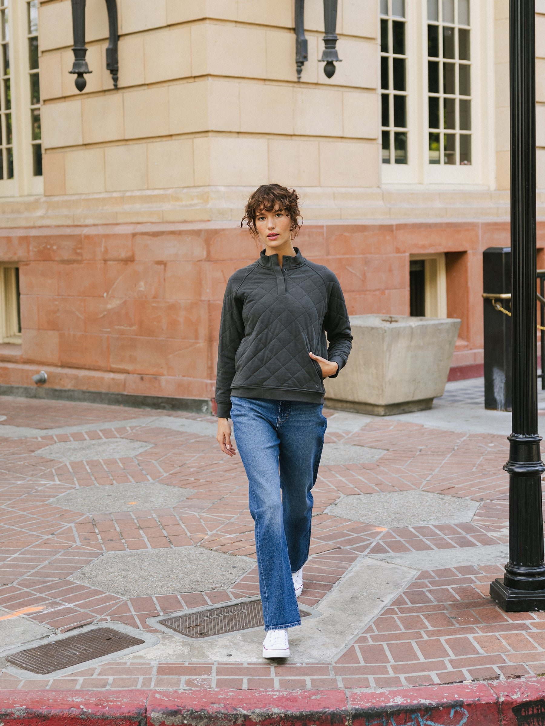 A woman with curly hair wearing a dark Cozy Earth Women's Ultra-Soft Bamboo Quilted Snap Pullover, blue jeans, and white sneakers walks along a brick-paved sidewalk. She has one hand in her pocket and is in front of a building with large windows. 