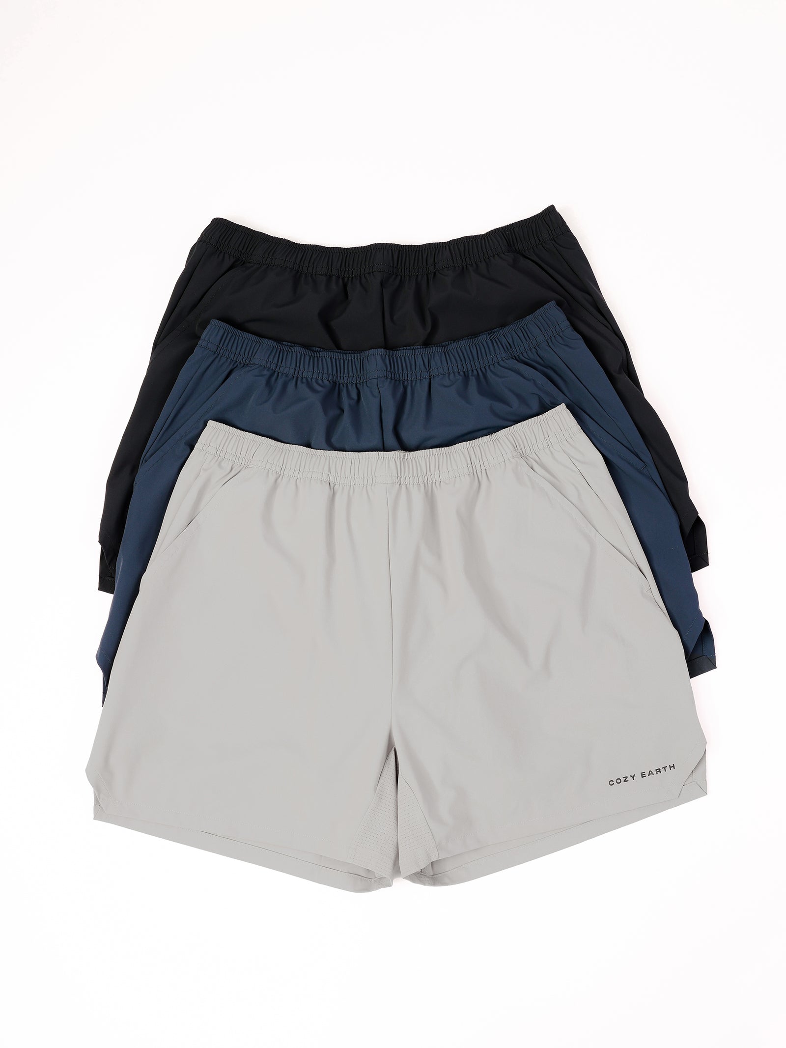 Three pairs of the Men's Performance Sleep Shorts by Cozy Earth are arranged in a stack. The top pair is black, the middle pair is dark blue, and the bottom pair is light gray. Each pair features an elastic waistband, and there is a "COZY EARTH" logo on the bottom left of the light gray shorts. 