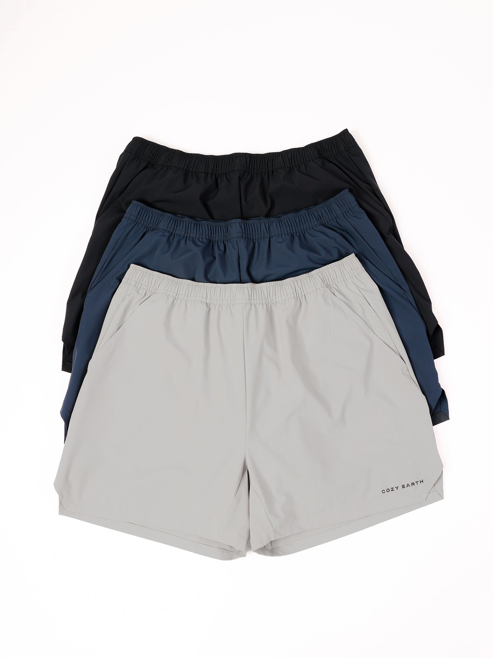 A stack of three Men's Performance Sleep Shorts from Cozy Earth in various colors is displayed. The top pair is black, the middle pair is dark blue, and the bottom pair is light gray, each featuring the "COZY EARTH" logo on the left leg. All shorts come with elastic waistbands. 