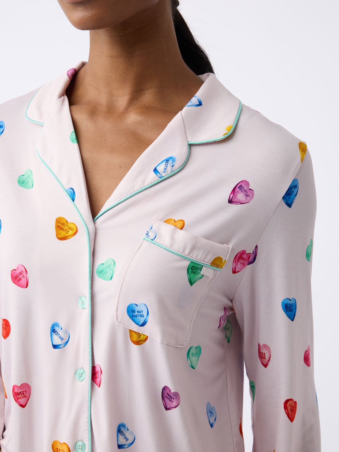 A woman wears the lilac-colored pajama shirt from the Women's Stretch-Knit Long Sleeve Bamboo Pajama Set by Cozy Earth, featuring colorful conversation hearts, buttons, a collar, chest pocket, and green piping. 